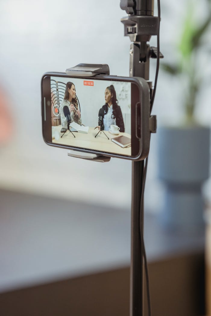 services-01 Young diverse women sitting at table with microphones and gadgets and talking while recording podcast on smartphone placed on tripod in studio